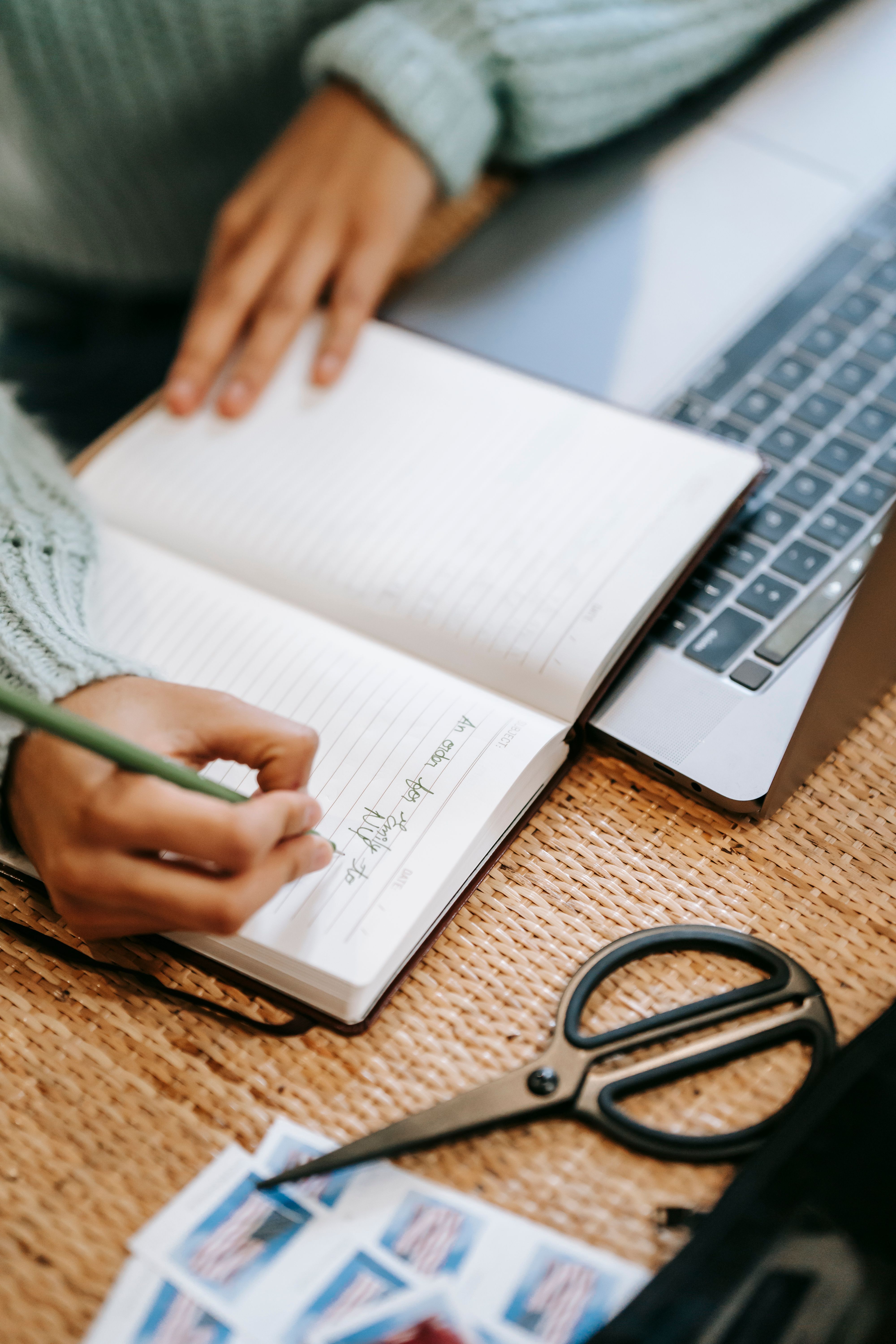 Hands taking notes beside a laptop on a woven tabletop